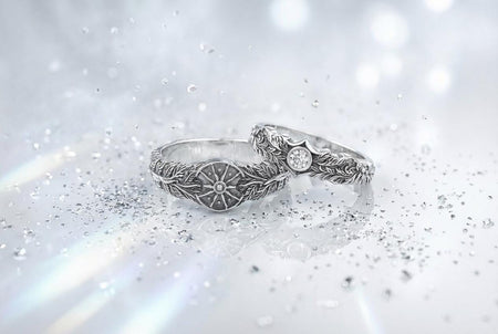 A macro studio shot of two matching antique-style sterling silver wedding rings resting on a sparkling, icy white surface. The wider men's band features a circular sunburst or shield motif with radiating lines. The narrower engagement ring features a matching sunburst centering a round white gemstone. Both bands are richly decorated with raised leaf and vine engravings, set against a dark oxidized background. __etsy:7975999301