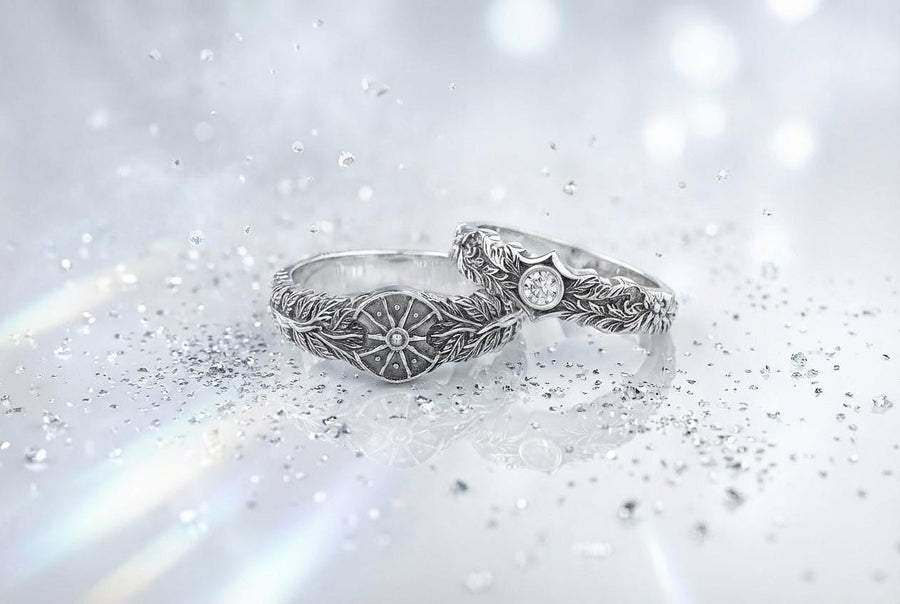 A macro studio shot of two matching antique-style sterling silver wedding rings resting on a sparkling, icy white surface. The wider men's band features a circular sunburst or shield motif with radiating lines. The narrower engagement ring features a matching sunburst centering a round white gemstone. Both bands are richly decorated with raised leaf and vine engravings, set against a dark oxidized background. __etsy:7975999301