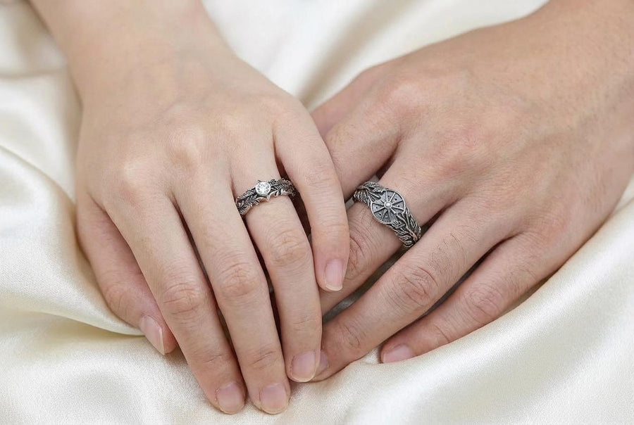 A macro studio shot of two matching antique-style sterling silver wedding rings resting on a sparkling, icy white surface. The wider men's band features a circular sunburst or shield motif with radiating lines. The narrower engagement ring features a matching sunburst centering a round white gemstone. Both bands are richly decorated with raised leaf and vine engravings, set against a dark oxidized background. __etsy:7975999301