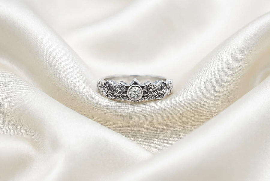A macro studio shot of two matching antique-style sterling silver wedding rings resting on a sparkling, icy white surface. The wider men's band features a circular sunburst or shield motif with radiating lines. The narrower engagement ring features a matching sunburst centering a round white gemstone. Both bands are richly decorated with raised leaf and vine engravings, set against a dark oxidized background. __etsy:7975999301
