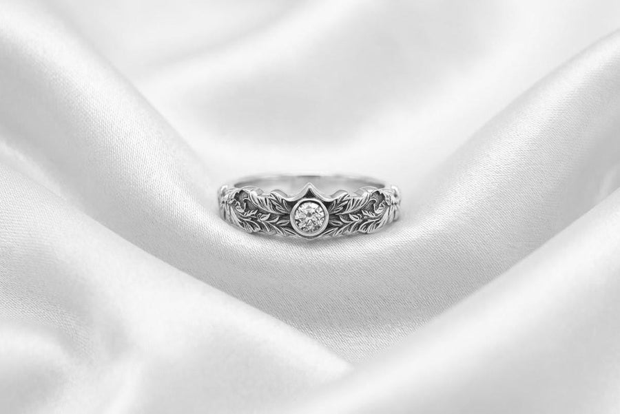 A macro studio shot of two matching antique-style sterling silver wedding rings resting on a sparkling, icy white surface. The wider men's band features a circular sunburst or shield motif with radiating lines. The narrower engagement ring features a matching sunburst centering a round white gemstone. Both bands are richly decorated with raised leaf and vine engravings, set against a dark oxidized background. __etsy:7975999301