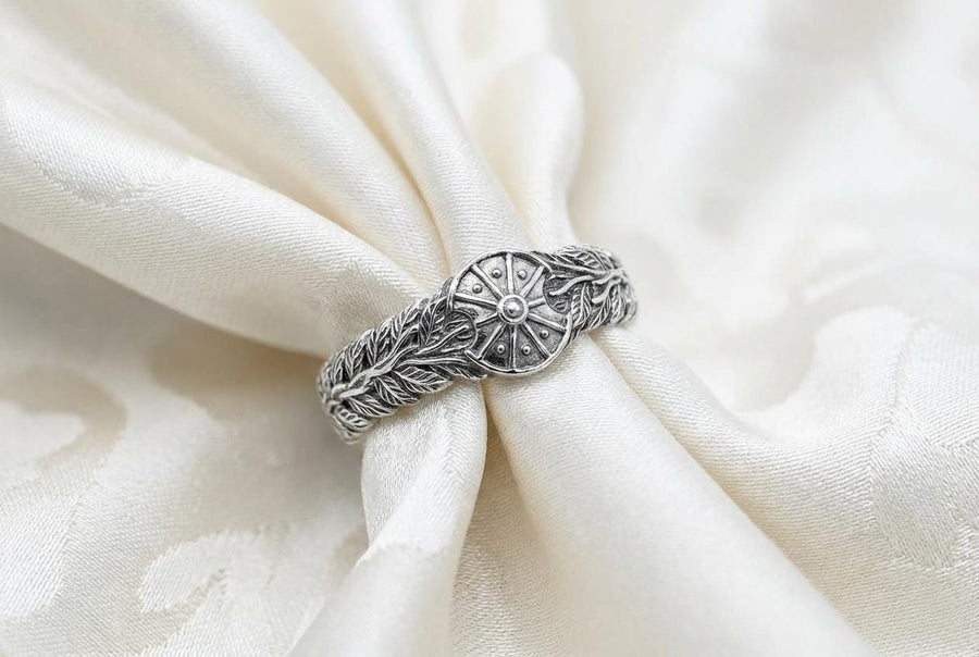 A macro studio shot of two matching antique-style sterling silver wedding rings resting on a sparkling, icy white surface. The wider men's band features a circular sunburst or shield motif with radiating lines. The narrower engagement ring features a matching sunburst centering a round white gemstone. Both bands are richly decorated with raised leaf and vine engravings, set against a dark oxidized background. __etsy:7975999301