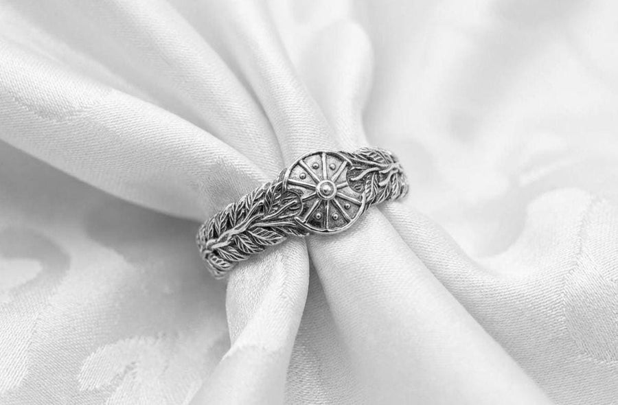 A macro studio shot of two matching antique-style sterling silver wedding rings resting on a sparkling, icy white surface. The wider men's band features a circular sunburst or shield motif with radiating lines. The narrower engagement ring features a matching sunburst centering a round white gemstone. Both bands are richly decorated with raised leaf and vine engravings, set against a dark oxidized background. __etsy:7975999301