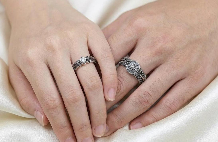 A macro studio shot of two matching antique-style sterling silver wedding rings resting on a sparkling, icy white surface. The wider men's band features a circular sunburst or shield motif with radiating lines. The narrower engagement ring features a matching sunburst centering a round white gemstone. Both bands are richly decorated with raised leaf and vine engravings, set against a dark oxidized background. __etsy:7975999301