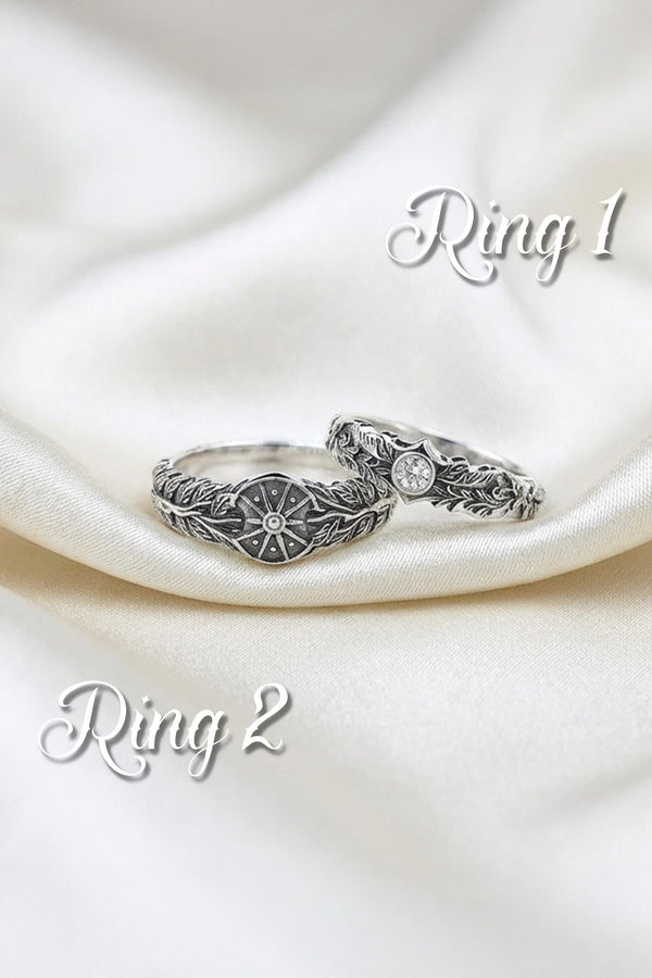 A macro studio shot of two matching antique-style sterling silver wedding rings resting on a sparkling, icy white surface. The wider men's band features a circular sunburst or shield motif with radiating lines. The narrower engagement ring features a matching sunburst centering a round white gemstone. Both bands are richly decorated with raised leaf and vine engravings, set against a dark oxidized background. __etsy:7975999301