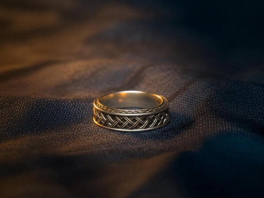 A cinematic studio shot of a wide sterling silver ring lying on a reflective golden surface. The band features a central "basket weave" pattern—diagonal strands of silver crossing over and under each other. The edges are framed by a distinct "rope" or "cable" twist border. The raised metal is polished silver, while the deep recesses are blackened to highlight the intricate weaving.
