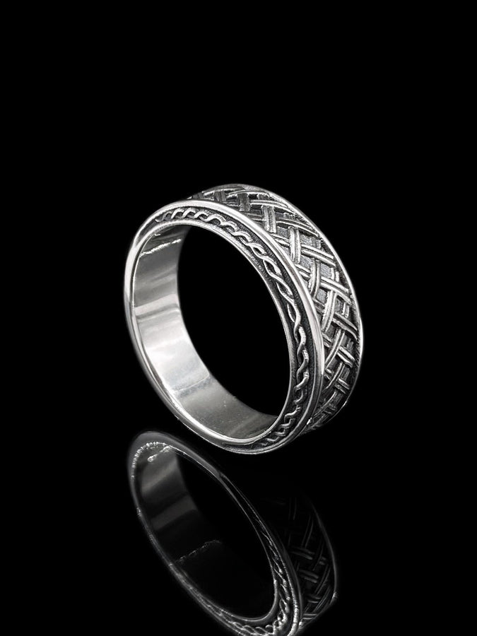 A cinematic studio shot of a wide sterling silver ring lying on a reflective golden surface. The band features a central "basket weave" pattern—diagonal strands of silver crossing over and under each other. The edges are framed by a distinct "rope" or "cable" twist border. The raised metal is polished silver, while the deep recesses are blackened to highlight the intricate weaving.