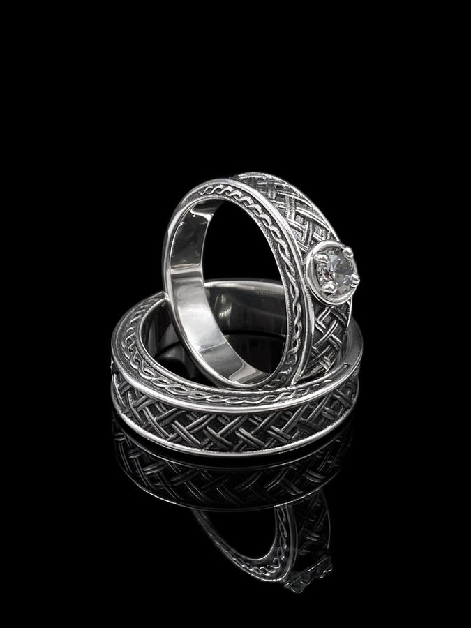 A cinematic studio shot of a wide sterling silver ring lying on a reflective golden surface. The band features a central "basket weave" pattern—diagonal strands of silver crossing over and under each other. The edges are framed by a distinct "rope" or "cable" twist border. The raised metal is polished silver, while the deep recesses are blackened to highlight the intricate weaving.