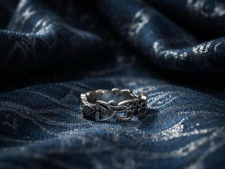 A cinematic close-up of a sterling silver ring lying on a reflective surface. The band features an "openwork" design, meaning the Celtic knot pattern is cut out, allowing light to pass through the loops. The silver strands weave over and under in a continuous infinity motif. The background is dark, but the ring is illuminated by a warm, golden mist.