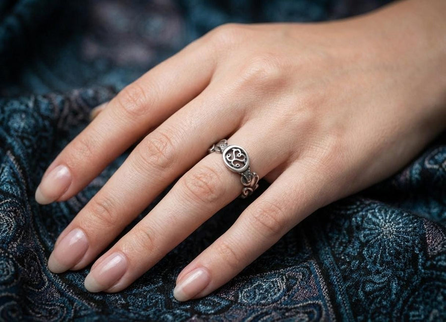 A cinematic macro shot of a sterling silver ring bathed in warm, golden light. The focal point is the central oval face, which encloses a raised relief design of two spirals meeting in the center to suggest a heart shape. The band shoulders feature textured knotwork that leads up to the centerpiece. The ring rests on a wet, reflective surface, creating a mirror image of the love knot below. (etsy image id: 7490345549)