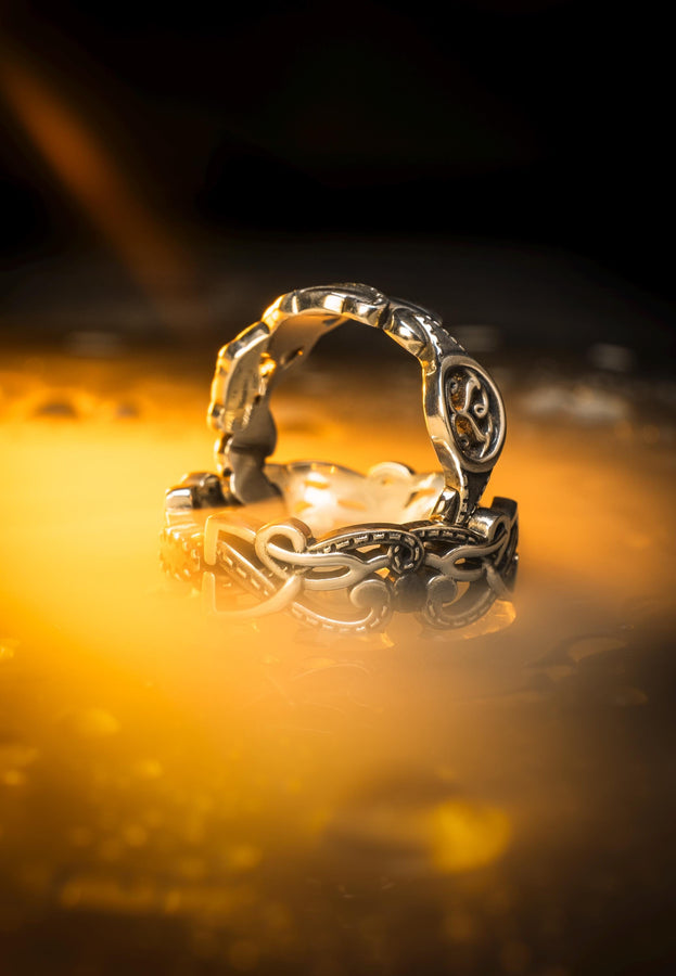 A cinematic macro shot of a sterling silver ring bathed in warm, golden light. The focal point is the central oval face, which encloses a raised relief design of two spirals meeting in the center to suggest a heart shape. The band shoulders feature textured knotwork that leads up to the centerpiece. The ring rests on a wet, reflective surface, creating a mirror image of the love knot below. (etsy image id: 7490345549)