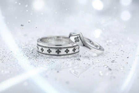 A high-resolution studio shot of a matching sterling silver wedding ring set resting on a sparkling, icy white surface. The set includes a wide unisex band and a delicate, V-shaped chevron ring. Both feature polished medieval quatrefoil motifs—four-lobed geometric shapes—set against a deeply textured, oxidized dark grey background that resembles crushed stone or stippled concrete. __etsy:7975881995