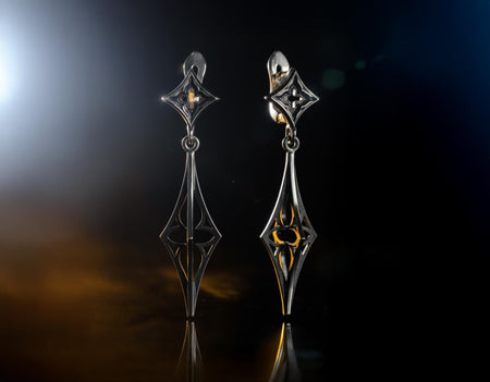 A cinematic studio shot of a pair of long, sharp sterling silver earrings standing upright. The structure consists of a top diamond-shaped stud and a bottom "stiletto" drop. The bottom piece is shaped like a slender needle or dagger, widening slightly in the middle to reveal a geometric cutout before tapering to a needle-sharp point. The silver is highly polished, gleaming against the darkness. (etsy image id: 7217885638)