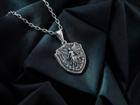 A highly detailed macro shot of a shield-shaped sterling silver pendant hanging from a silver chain. The center features a three-dimensional bas-relief of Nike, the winged Greek goddess of victory, holding a laurel wreath. She stands gracefully against a deeply oxidized background depicting classical Greek temple architecture with fluted columns. The raised silver highlights contrast with the dark antiqued recesses. __etsy:8017759430