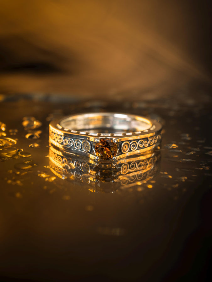 A detailed macro shot of a sterling silver engagement ring. The focal point is a round, honey-colored gemstone (Citrine or Madeira Topaz) set flush into the band. The shank is decorated with a continuous relief pattern of spirals and circles, mimicking the decorative robes found in Gustav Klimt's "Golden Phase" paintings. The silver features an oxidized finish, creating a dark background that makes the polished spirals pop. (etsy image id: 7142785932)