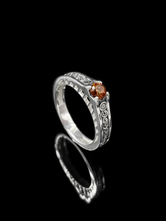 A detailed macro shot of a sterling silver engagement ring. The focal point is a round, honey-colored gemstone (Citrine or Madeira Topaz) set flush into the band. The shank is decorated with a continuous relief pattern of spirals and circles, mimicking the decorative robes found in Gustav Klimt's "Golden Phase" paintings. The silver features an oxidized finish, creating a dark background that makes the polished spirals pop. (etsy image id: 7142785932)