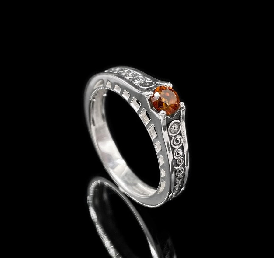 A detailed macro shot of a sterling silver engagement ring. The focal point is a round, honey-colored gemstone (Citrine or Madeira Topaz) set flush into the band. The shank is decorated with a continuous relief pattern of spirals and circles, mimicking the decorative robes found in Gustav Klimt's "Golden Phase" paintings. The silver features an oxidized finish, creating a dark background that makes the polished spirals pop. (etsy image id: 7142785932)