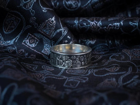 A detailed profile shot of a wide sterling silver ring standing on a reflective surface. The band features a raised relief carving of the figures from Gustav Klimt's painting "The Kiss." To the left of the couple are sharp, vertical rectangular blocks. To the right are swirling circular spirals. The background of the carving is darkened with oxidation to contrast with the polished silver figures. __etsy:7442609320