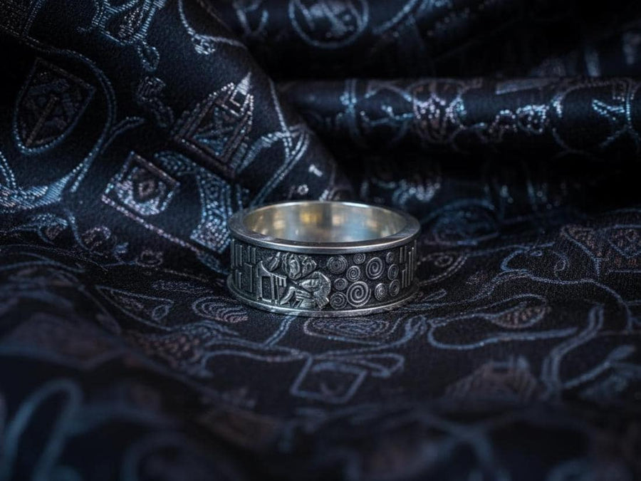 A detailed profile shot of a wide sterling silver ring standing on a reflective surface. The band features a raised relief carving of the figures from Gustav Klimt's painting "The Kiss." To the left of the couple are sharp, vertical rectangular blocks. To the right are swirling circular spirals. The background of the carving is darkened with oxidation to contrast with the polished silver figures. __etsy:7442609320