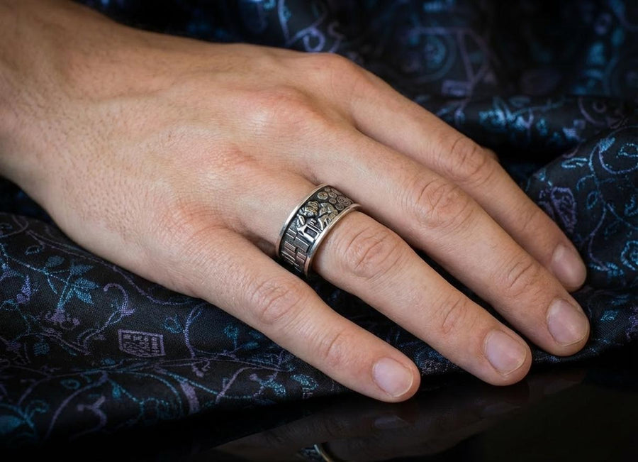 A detailed profile shot of a wide sterling silver ring standing on a reflective surface. The band features a raised relief carving of the figures from Gustav Klimt's painting "The Kiss." To the left of the couple are sharp, vertical rectangular blocks. To the right are swirling circular spirals. The background of the carving is darkened with oxidation to contrast with the polished silver figures. __etsy:7442609320
