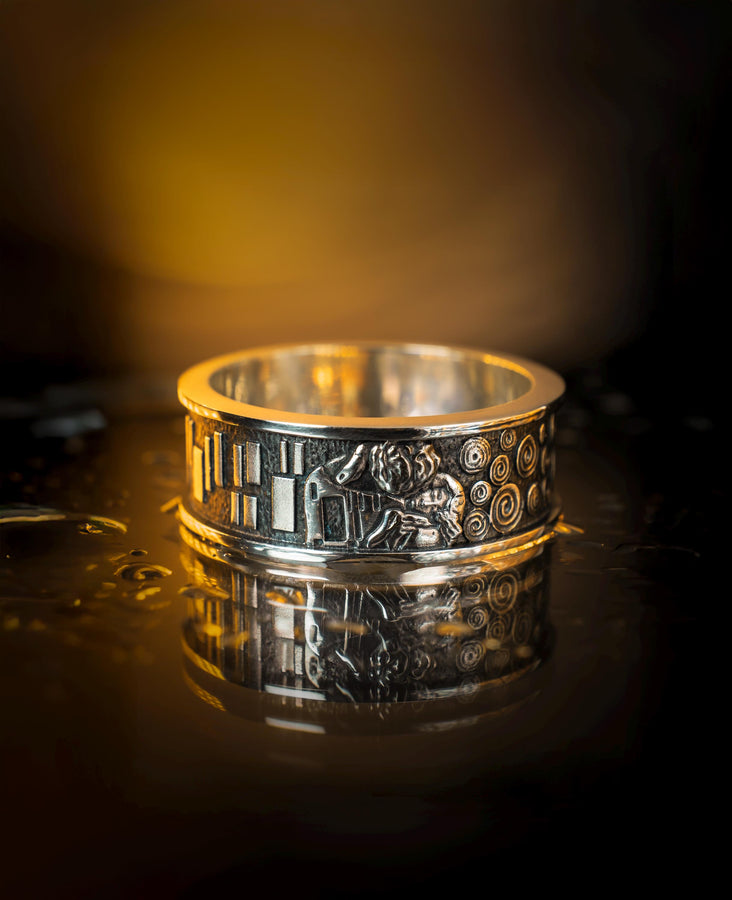 A detailed profile shot of a wide sterling silver ring standing on a reflective surface. The band features a raised relief carving of the figures from Gustav Klimt's painting "The Kiss." To the left of the couple are sharp, vertical rectangular blocks. To the right are swirling circular spirals. The background of the carving is darkened with oxidation to contrast with the polished silver figures. __etsy:7442609320