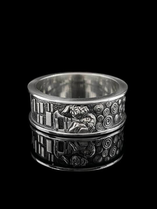 A detailed profile shot of a wide sterling silver ring standing on a reflective surface. The band features a raised relief carving of the figures from Gustav Klimt's painting "The Kiss." To the left of the couple are sharp, vertical rectangular blocks. To the right are swirling circular spirals. The background of the carving is darkened with oxidation to contrast with the polished silver figures. __etsy:7442609320