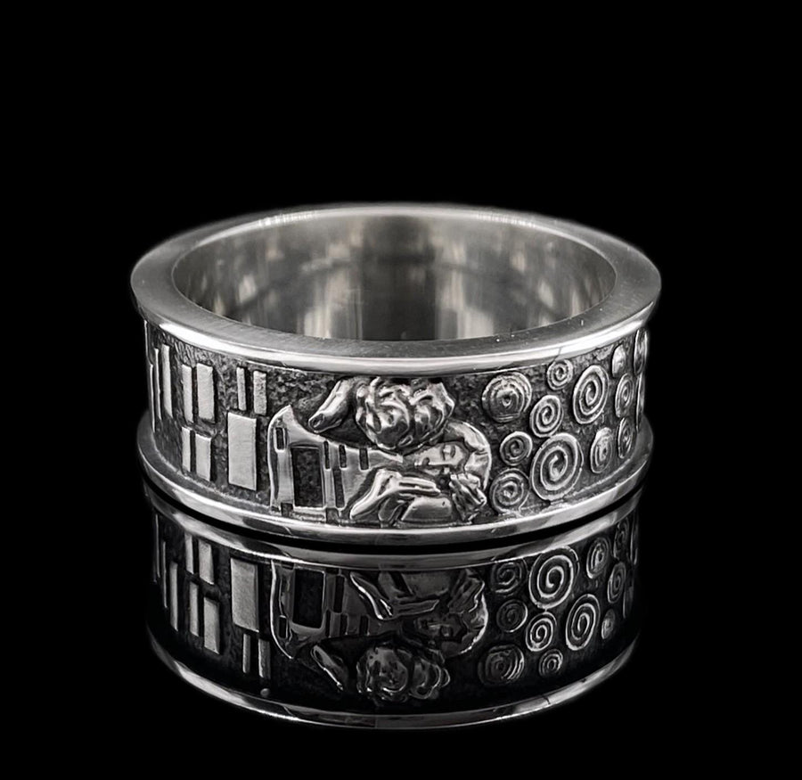 A detailed profile shot of a wide sterling silver ring standing on a reflective surface. The band features a raised relief carving of the figures from Gustav Klimt's painting "The Kiss." To the left of the couple are sharp, vertical rectangular blocks. To the right are swirling circular spirals. The background of the carving is darkened with oxidation to contrast with the polished silver figures. __etsy:7442609320