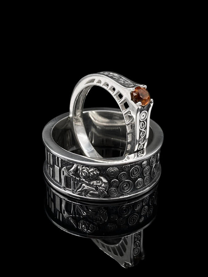 A detailed profile shot of a wide sterling silver ring standing on a reflective surface. The band features a raised relief carving of the figures from Gustav Klimt's painting "The Kiss." To the left of the couple are sharp, vertical rectangular blocks. To the right are swirling circular spirals. The background of the carving is darkened with oxidation to contrast with the polished silver figures. __etsy:7442609320
