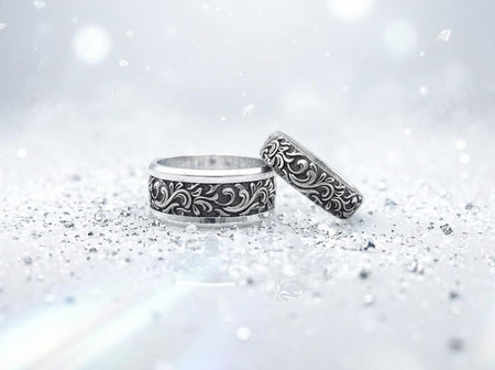 A close-up studio shot of a matching organic gothic wedding band set in sterling silver, resting on a dark, reflective surface. The rings feature a continuous, high-relief pattern of intricate sterling silver vine patterns and swirling leaves. The recessed areas are heavily oxidized to a dark matte black, creating a high-contrast shadow effect that makes the polished organic scrollwork stand out brilliantly against the dark background. __etsy:8017806924