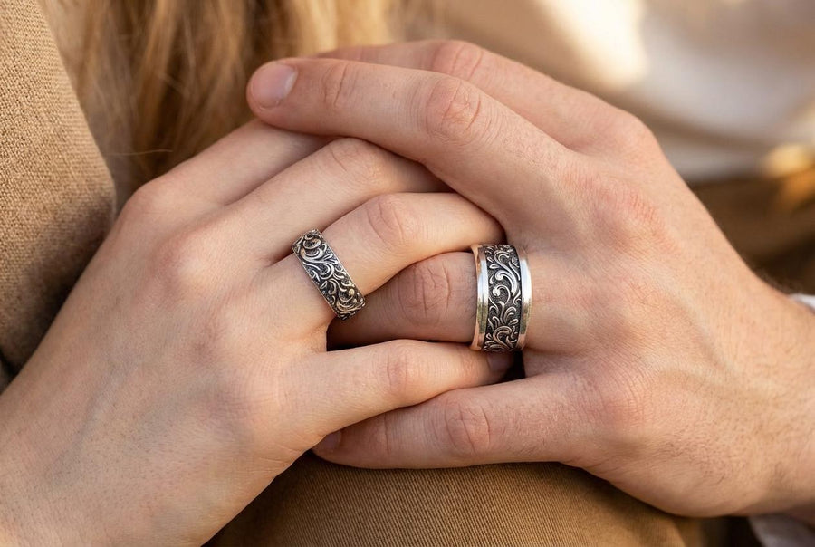 A close-up studio shot of a matching organic gothic wedding band set in sterling silver, resting on a dark, reflective surface. The rings feature a continuous, high-relief pattern of intricate sterling silver vine patterns and swirling leaves. The recessed areas are heavily oxidized to a dark matte black, creating a high-contrast shadow effect that makes the polished organic scrollwork stand out brilliantly against the dark background. __etsy:8017806924