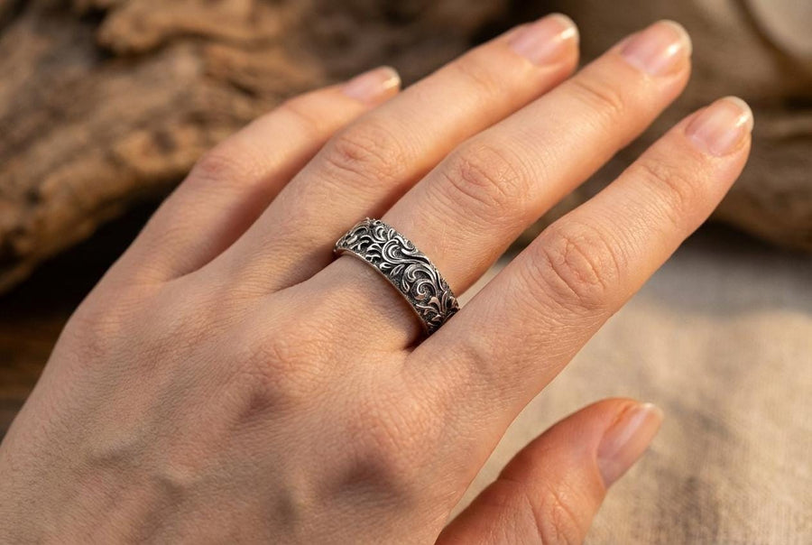 A close-up studio shot of a matching organic gothic wedding band set in sterling silver, resting on a dark, reflective surface. The rings feature a continuous, high-relief pattern of intricate sterling silver vine patterns and swirling leaves. The recessed areas are heavily oxidized to a dark matte black, creating a high-contrast shadow effect that makes the polished organic scrollwork stand out brilliantly against the dark background. __etsy:8017806924