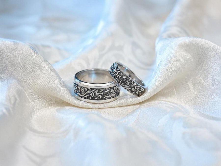 A close-up studio shot of a matching organic gothic wedding band set in sterling silver, resting on a dark, reflective surface. The rings feature a continuous, high-relief pattern of intricate sterling silver vine patterns and swirling leaves. The recessed areas are heavily oxidized to a dark matte black, creating a high-contrast shadow effect that makes the polished organic scrollwork stand out brilliantly against the dark background. __etsy:8017806924
