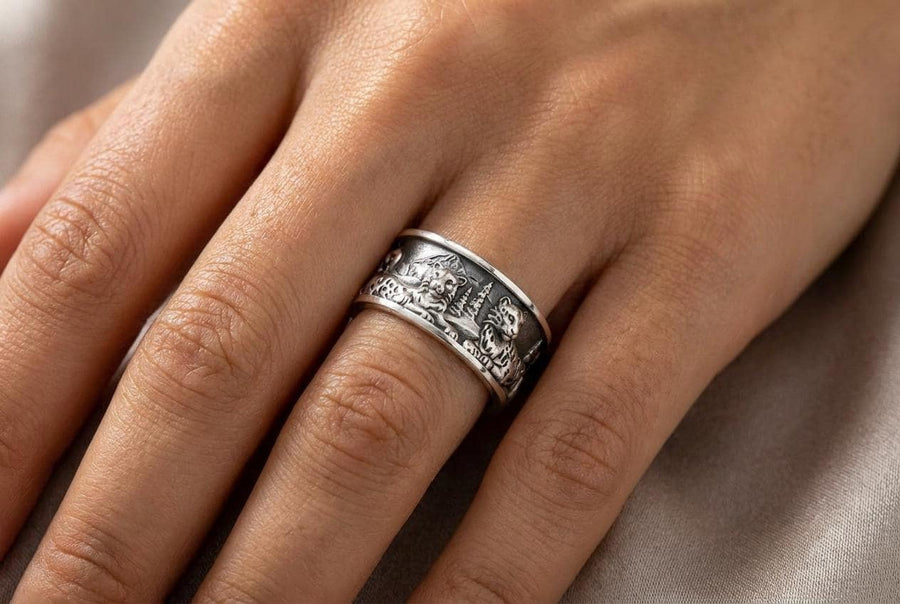 A close-up studio shot of a wide, open-back sterling silver cuff ring resting on a black reflective surface. The band features a detailed, three-dimensional bas-relief carving of a mountain landscape. In the foreground, two realistic snow leopards lie side by side among textured pine trees and rocky peaks. The 925 silver is heavily oxidized in the recesses to highlight the intricate fur spots, trees, and mountainous terrain against the polished r __etsy:7979063548