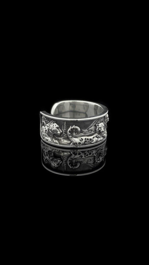 A close-up studio shot of a wide, open-back sterling silver cuff ring resting on a black reflective surface. The band features a detailed, three-dimensional bas-relief carving of a mountain landscape. In the foreground, two realistic snow leopards lie side by side among textured pine trees and rocky peaks. The 925 silver is heavily oxidized in the recesses to highlight the intricate fur spots, trees, and mountainous terrain against the polished r __etsy:7979063548