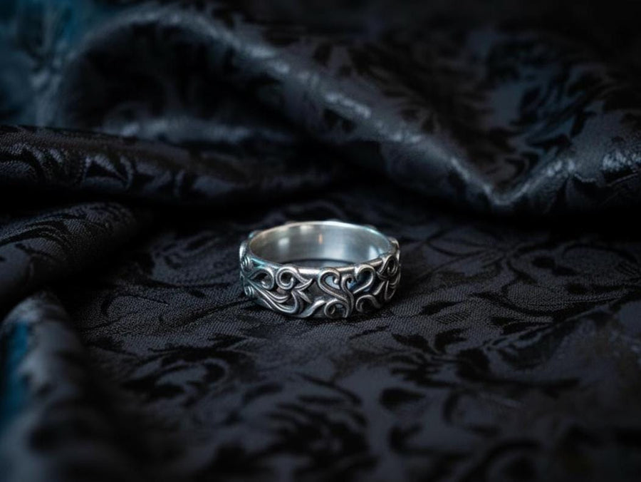 A detailed close-up of a wide sterling silver ring resting on a reflective surface. The band is covered in a deep-relief filigree pattern consisting of interlocking scrolls, vines, and subtle heart shapes. The raised designs are polished bright silver, while the recessed background is heavily oxidized to black, highlighting the complexity of the Baroque motif.