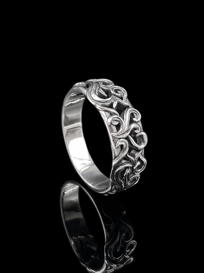 A detailed close-up of a wide sterling silver ring resting on a reflective surface. The band is covered in a deep-relief filigree pattern consisting of interlocking scrolls, vines, and subtle heart shapes. The raised designs are polished bright silver, while the recessed background is heavily oxidized to black, highlighting the complexity of the Baroque motif.