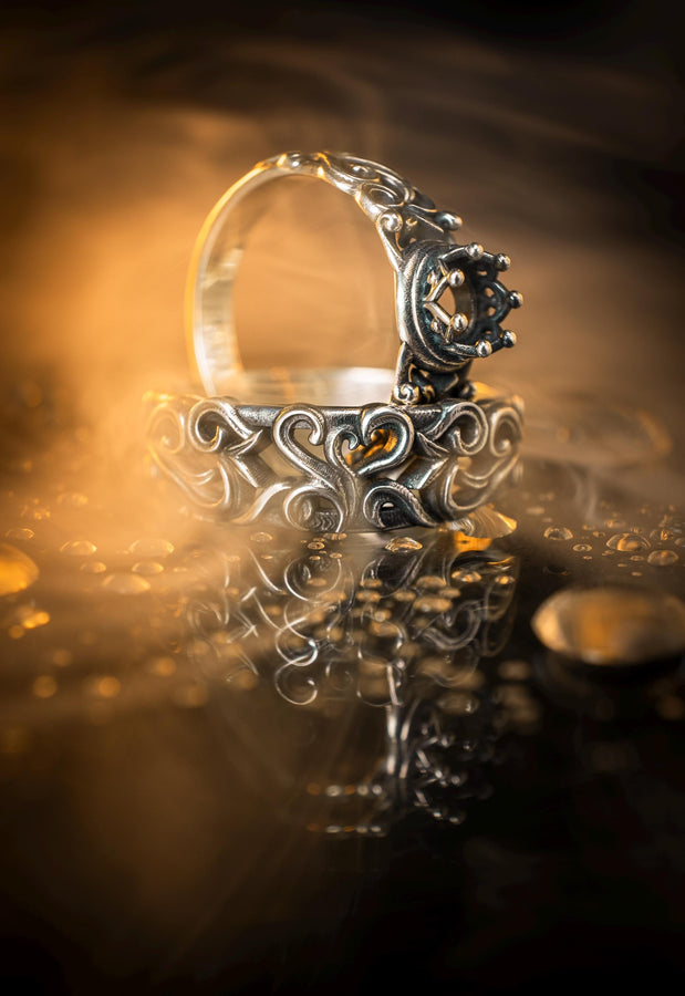 A detailed close-up of a wide sterling silver ring resting on a reflective surface. The band is covered in a deep-relief filigree pattern consisting of interlocking scrolls, vines, and subtle heart shapes. The raised designs are polished bright silver, while the recessed background is heavily oxidized to black, highlighting the complexity of the Baroque motif.