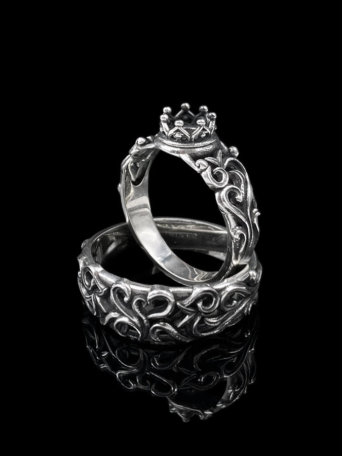 A detailed close-up of a wide sterling silver ring resting on a reflective surface. The band is covered in a deep-relief filigree pattern consisting of interlocking scrolls, vines, and subtle heart shapes. The raised designs are polished bright silver, while the recessed background is heavily oxidized to black, highlighting the complexity of the Baroque motif.