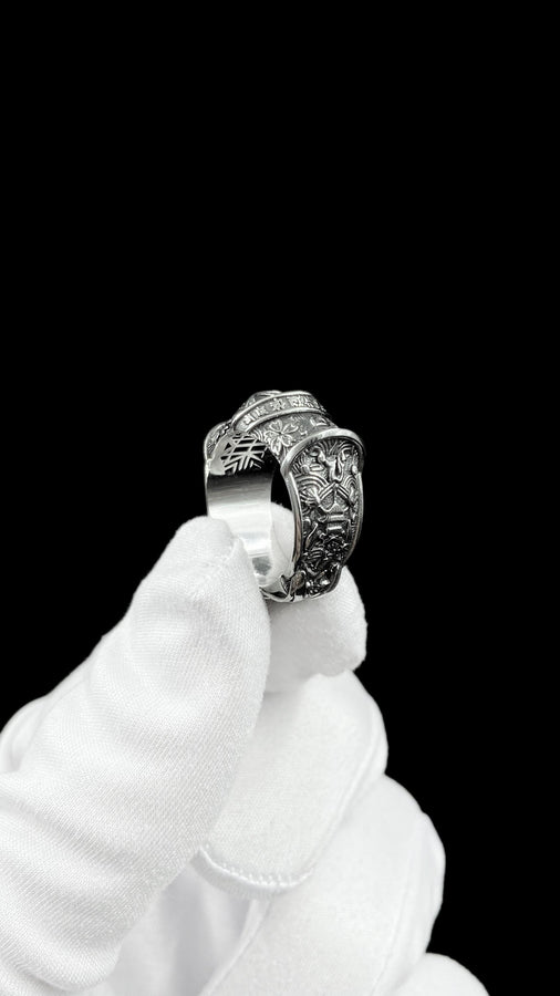 A close-up studio photograph of a wide sterling silver ring designed to look like folded fabric. The overlapping top mimics an Obi, the traditional Japanese kimono sash. The band features highly detailed relief carvings of Sakura (cherry blossoms) and Seigaiha (concentric ocean waves) patterns. A raised ribbon section bears engraved Japanese Kanji script. The silver is heavily oxidized in the recesses to highlight the intricate textures against t (etsy image id: 7831393878)