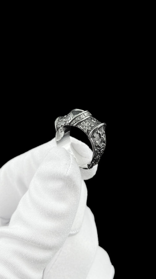 A close-up studio photograph of a wide sterling silver ring designed to look like folded fabric. The overlapping top mimics an Obi, the traditional Japanese kimono sash. The band features highly detailed relief carvings of Sakura (cherry blossoms) and Seigaiha (concentric ocean waves) patterns. A raised ribbon section bears engraved Japanese Kanji script. The silver is heavily oxidized in the recesses to highlight the intricate textures against t (etsy image id: 7831393878)