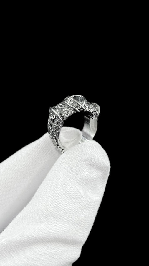 A close-up studio photograph of a wide sterling silver ring designed to look like folded fabric. The overlapping top mimics an Obi, the traditional Japanese kimono sash. The band features highly detailed relief carvings of Sakura (cherry blossoms) and Seigaiha (concentric ocean waves) patterns. A raised ribbon section bears engraved Japanese Kanji script. The silver is heavily oxidized in the recesses to highlight the intricate textures against t (etsy image id: 7831393878)