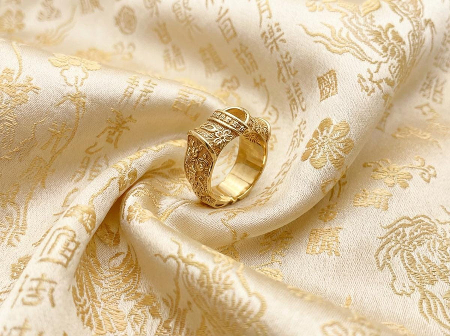 A close-up studio photograph of a wide sterling silver ring designed to look like folded fabric. The overlapping top mimics an Obi, the traditional Japanese kimono sash. The band features highly detailed relief carvings of Sakura (cherry blossoms) and Seigaiha (concentric ocean waves) patterns. A raised ribbon section bears engraved Japanese Kanji script. The silver is heavily oxidized in the recesses to highlight the intricate textures against t (etsy image id: 7831393878)