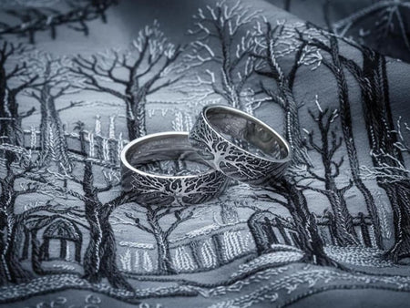 A clean studio shot of two matching sterling silver rings resting on a slate grey surface. Both bands feature a continuous panoramic design of a tree with spreading branches and deep roots. The tree motif is polished silver, which contrasts sharply with the textured, oxidized grey background that resembles tree bark or drifting soil. __etsy:7444576854