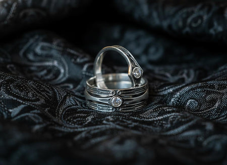 A high-contrast studio shot of a two-piece sterling silver ring set. The foreground features a wide band lying flat, set with a round clear gemstone. Flanking the stone are horizontal, grooved lines resembling arrows or spears. Standing behind it is a narrower engagement-style ring with a matching "lance-tip" shank leading to a central bezel-set white stone. The background is a textured, dark fabric. __etsy:7441122056