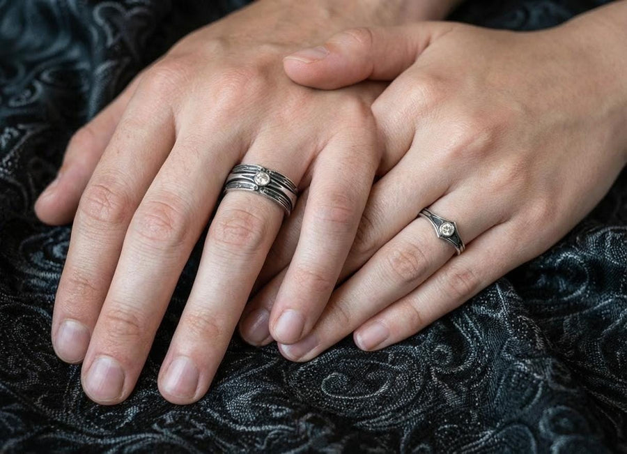 A high-contrast studio shot of a two-piece sterling silver ring set. The foreground features a wide band lying flat, set with a round clear gemstone. Flanking the stone are horizontal, grooved lines resembling arrows or spears. Standing behind it is a narrower engagement-style ring with a matching "lance-tip" shank leading to a central bezel-set white stone. The background is a textured, dark fabric. __etsy:7441122056