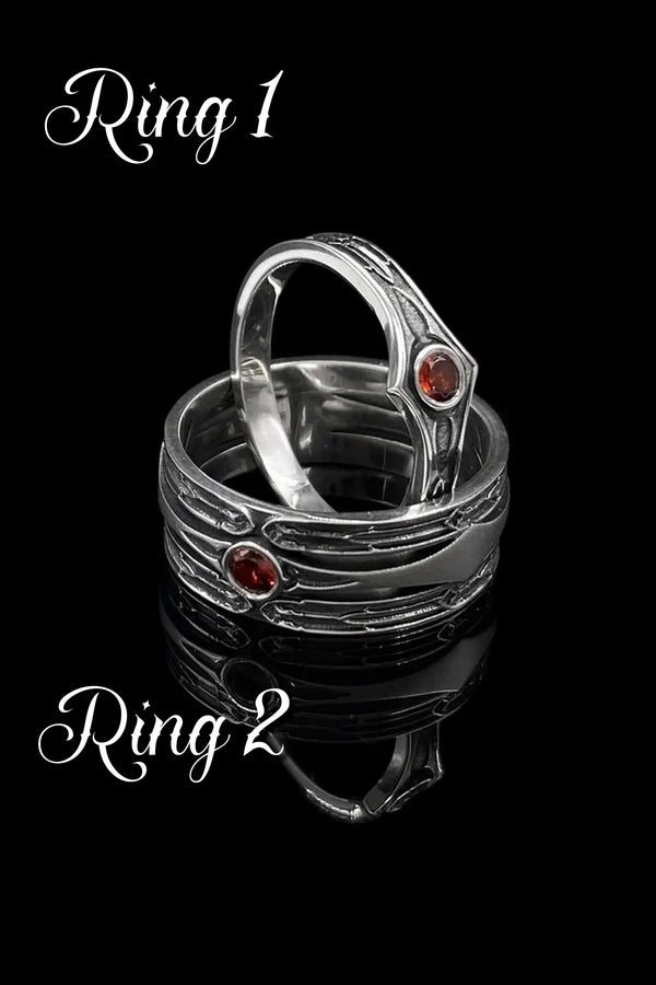 A high-contrast studio shot of a two-piece sterling silver ring set. The foreground features a wide band lying flat, set with a round clear gemstone. Flanking the stone are horizontal, grooved lines resembling arrows or spears. Standing behind it is a narrower engagement-style ring with a matching "lance-tip" shank leading to a central bezel-set white stone. The background is a textured, dark fabric. __etsy:7441122056