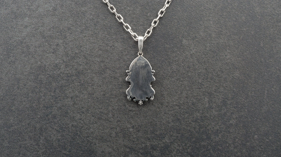 A detailed studio shot of a sterling silver pendant resting on a dark, textured stone surface. The design features a realistic human face in the center, but the forehead and eyes are obscured by a grid of Gothic window tracery (vertical bars and trefoils). The chin is framed by swirling Art Nouveau-style acanthus leaves. The silver is heavily oxidized, giving the face the appearance of a weathered stone statue. (etsy image id: 7448678222)
