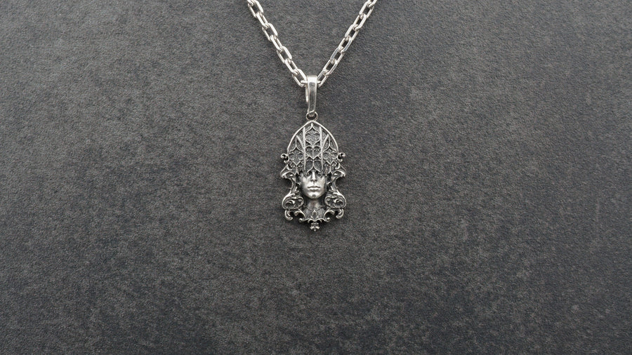 A detailed studio shot of a sterling silver pendant resting on a dark, textured stone surface. The design features a realistic human face in the center, but the forehead and eyes are obscured by a grid of Gothic window tracery (vertical bars and trefoils). The chin is framed by swirling Art Nouveau-style acanthus leaves. The silver is heavily oxidized, giving the face the appearance of a weathered stone statue. (etsy image id: 7448678222)