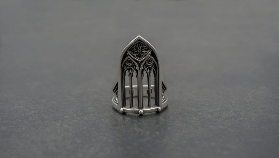 A clean studio shot of a tall, architectural sterling silver ring standing on a matte grey surface. The ring creates the silhouette of a Gothic "Lancet Window." It features three vertical openings separated by slender bars (mullions), topped by an intricate geometric tracery design within the pointed arch. The metal is matte silver with dark oxidation in the corners. (etsy image id: 7500328125)