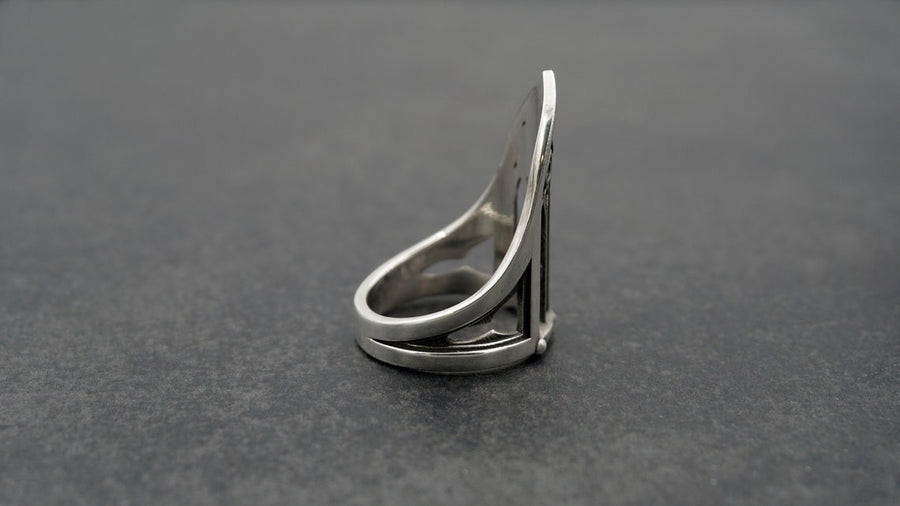 A clean studio shot of a tall, architectural sterling silver ring standing on a matte grey surface. The ring creates the silhouette of a Gothic "Lancet Window." It features three vertical openings separated by slender bars (mullions), topped by an intricate geometric tracery design within the pointed arch. The metal is matte silver with dark oxidation in the corners. (etsy image id: 7500328125)